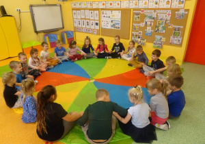 Grupa dzieci siedzi na brzegu chusty animacyjnej w siadzie skrzyżnym z dłońmi położonymi na kolanach kolegi bądź koleżanki siedzących po prawej i lewej stronie.